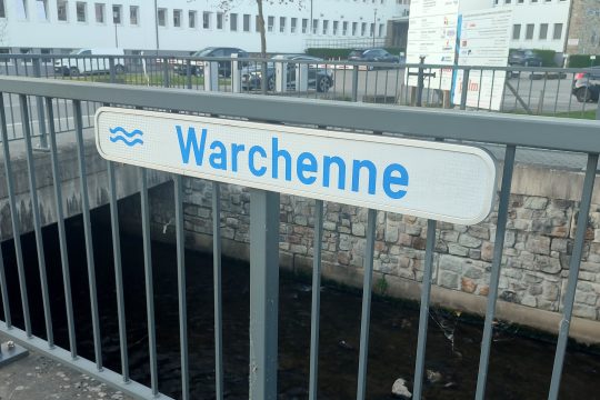 Schild mit der Beschriftung "Warchenne" an einem Geländer am Bachlauf
