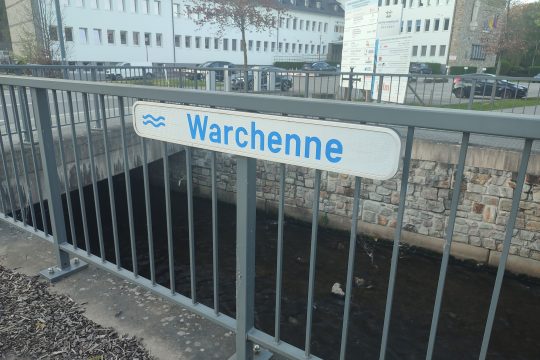 Schild mit der Beschriftung "Warchenne" ist an einem Geländer am Bachlauf befestigt