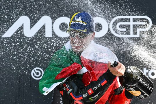 Marco Bezzecchi steht auf dem Podium, mit einer italienischen Flagge um den Hals, er macht eine Champagnerdusche