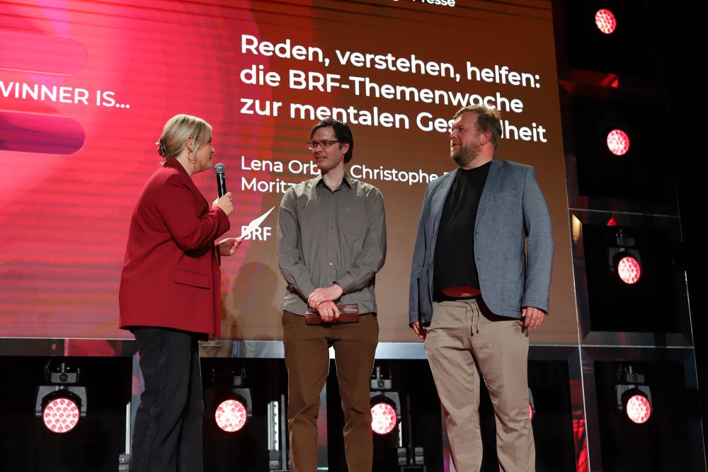 Die BRF-Journalisten Moritz Korff und Christophe Ramjoie stehen auf der Bühne und werden von Moderatorin Ophélie Fontana interviewt