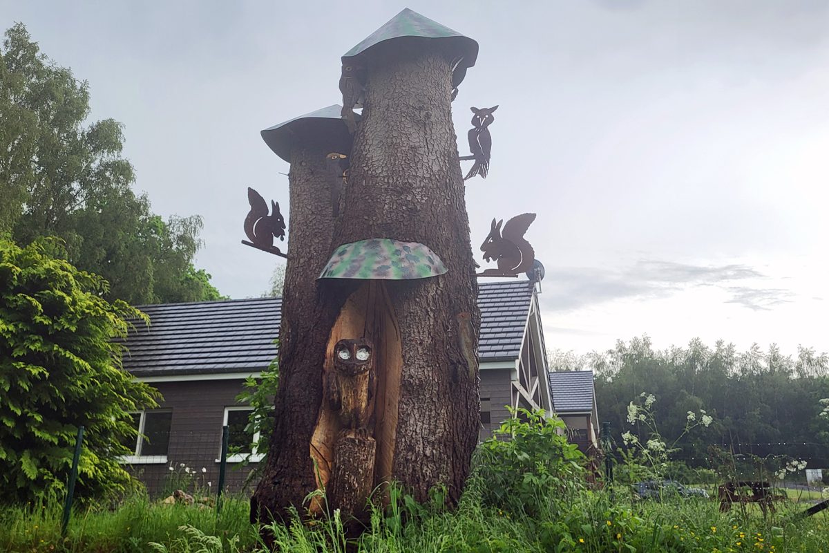 Eine Kunstfigur bestehend aus Baumstämmen, die an ein Schloss erinnern mit metallenen Silhouettenfiguren, die Eulen und andere Waldtiere darstellen