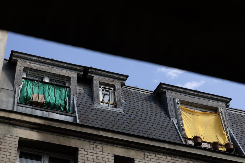 Dachgeschoss eines Pariser Wohngebäudes, an einigen Fenstern hängen Tücher als improvisierter Sonnenschutz