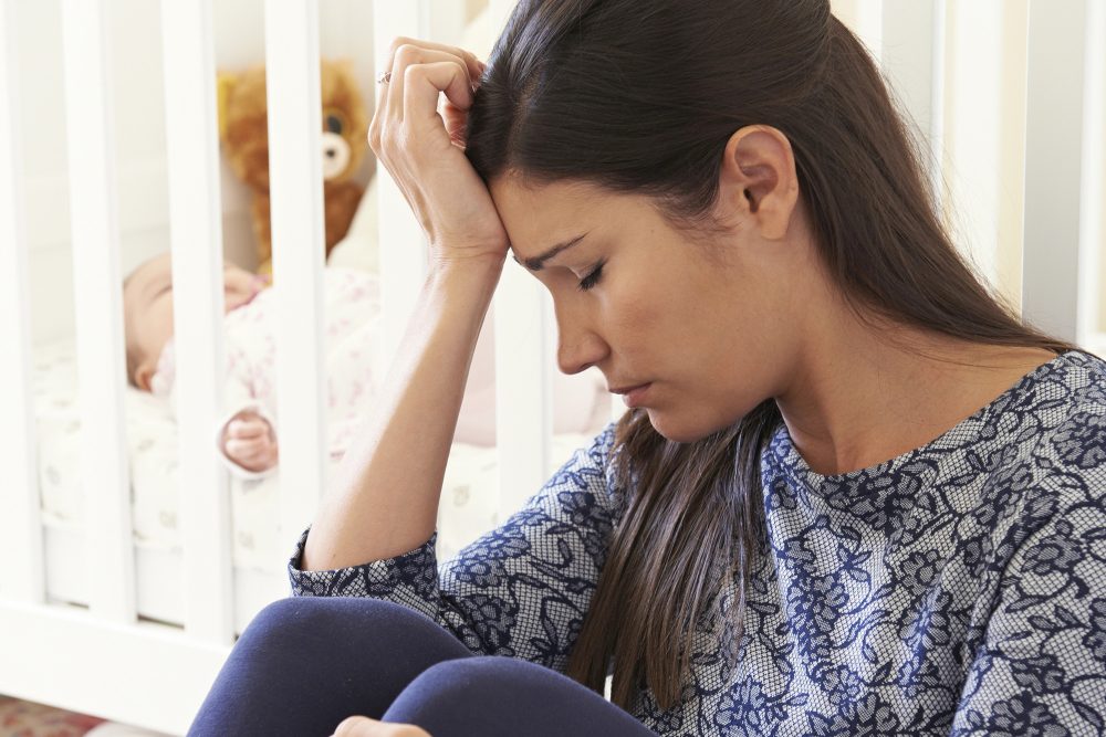 Erschöpfte Mutter mit Baby im Gitterbett im Hintergrund