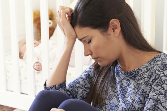Erschöpfte Mutter mit Baby im Gitterbett im Hintergrund
