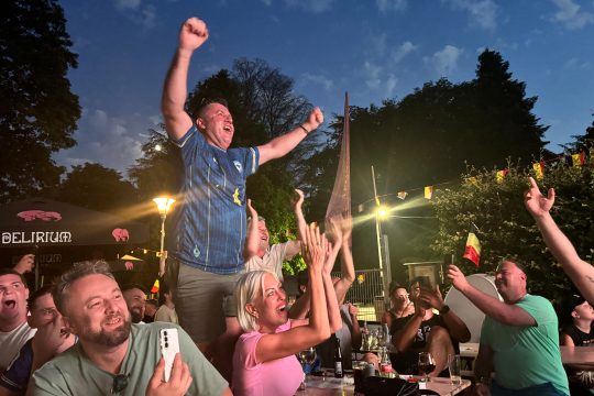 Menschen sitzen beim Public Viewing an Tischen, ein Mann hat sich auf eine Bank gestellt und reckt jubelnd die Arme in die Höhe