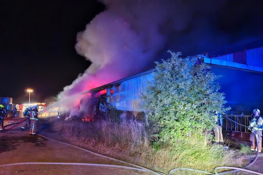 Feuerwehrleute löschen die brennende Lagerhalle, dichter Rauch steigt auf