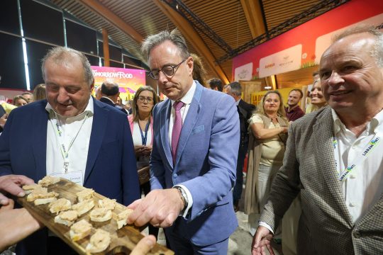 Premier De Wever nimmt ein belegtes Brot in die Hand, das ihm auf einem Holzbrett angeboten wird