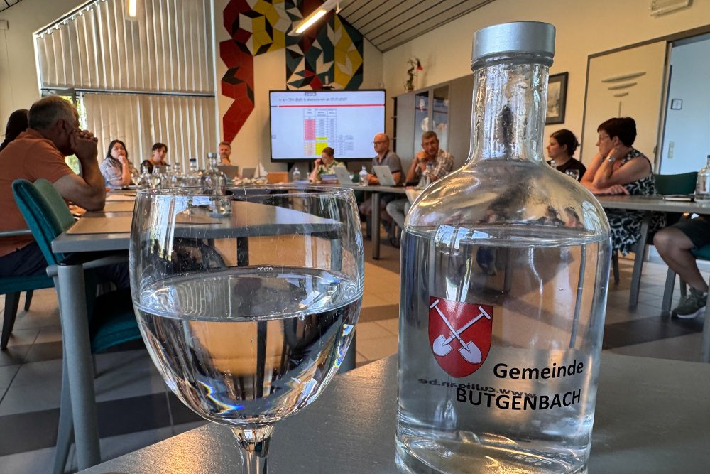 Wasserflasche mit der Aufschrift "Gemeinde Bütgenbach" und dem Gemeindewappen, daneben ein halb gefülltes Glas - im Hintergrund tagt der Gemeinderat
