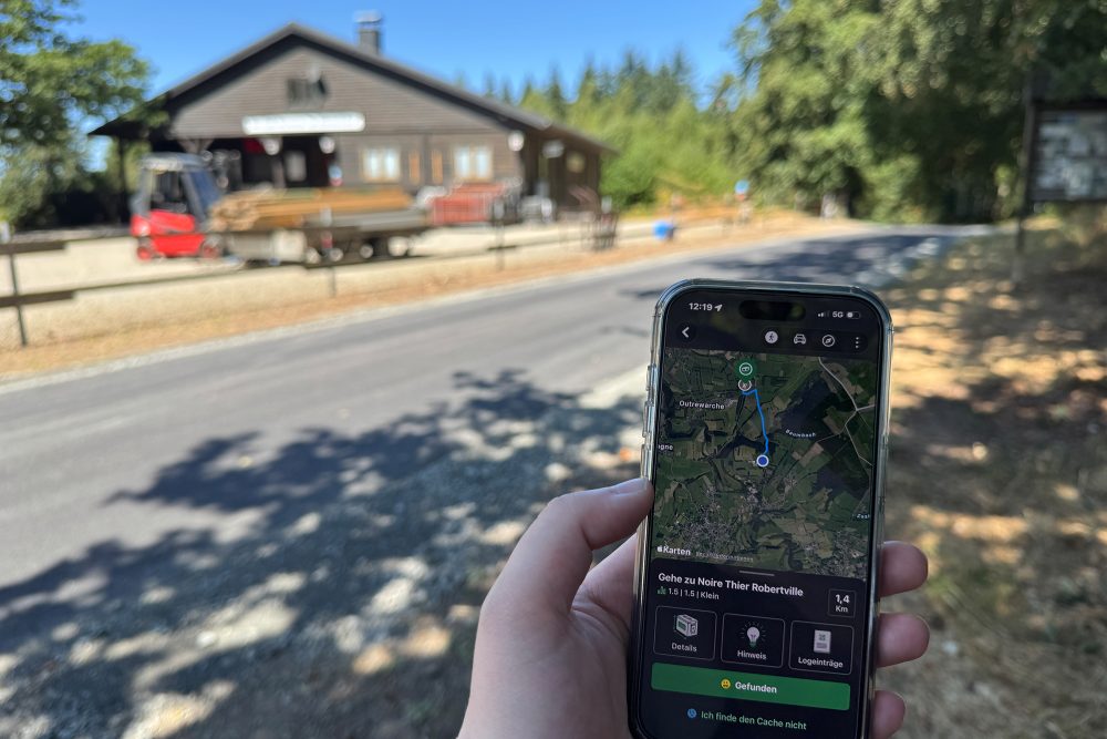 In einer Hand wird ein Smartphone gehalten, auf dem die Geocaching-App offen ist - im Hintergrund sieht man die Skihütte Himmelchen in Weywertz