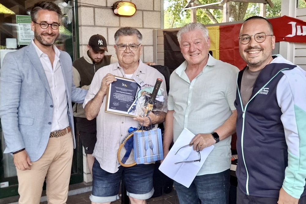 Raymond Schmetz hält eine gerahmte Urkunde in den Händen, einen Tennisschläger, eine Weinflasche und eine Geschenktüte - um ihn herum sind Bürgermeister Daniel Hilligsmann, Schöffe Björn Klinkenberg und eine weitere Person