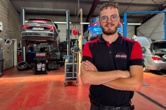 Kfz-Mechatroniker John Wiesemes in der Garage, im Hintergrund ein Auto auf der Hebebühne