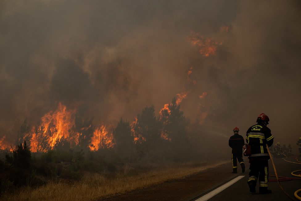 Wald steht in Flammen, daneben eine Straße, auf der Feuerwehrleute stehen, und versuchen zu löschen