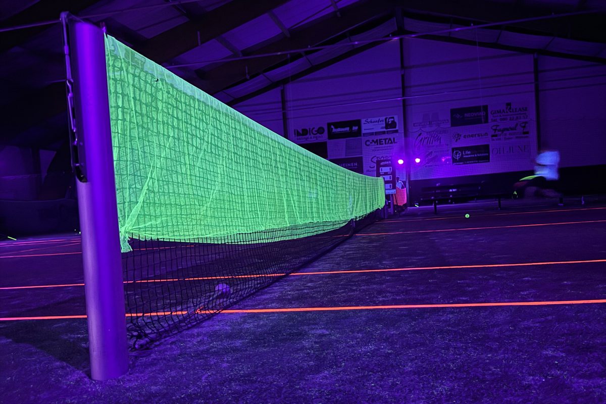 Das Tennisnetz leuchtet im Schwarzlicht neongelb, die Platzbegrenzungen leuchten rot