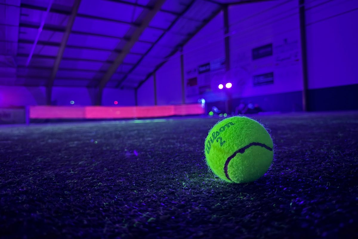 Ein leuchtend neongelber Tennisball auf dem Boden, im Hintergrund ein rosa leuchtendes Tennisnetz