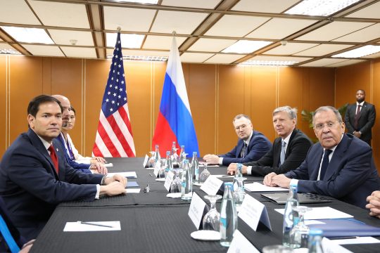 Marco Rubio und Sergej Lawrow sowie weitere Männer sitzen an einem Konferenztisch, im Hintergrund sind die US-amerikanische und die russische Flagge aufgestellt