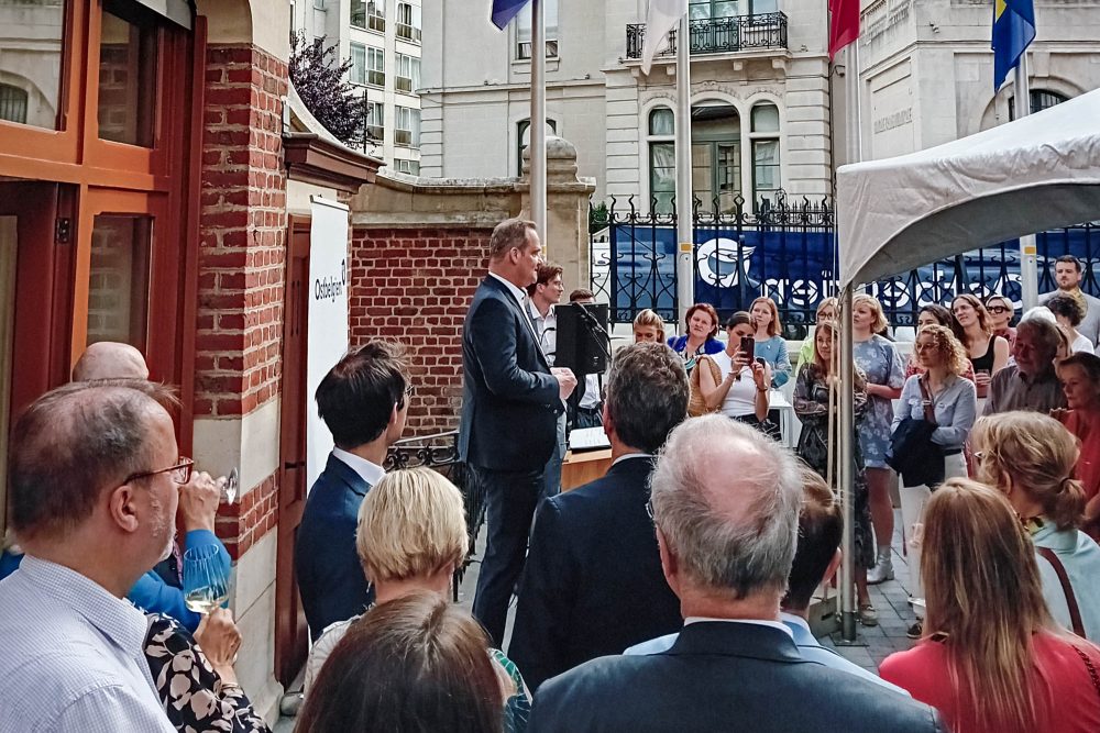 Ministerpräsident Paasch spricht im Hof zu den Gästen. Im Hintergrund die Flagge der EU, die belgische Fahne und die Fahnde der Deutschsprachigen Gemeinschaft