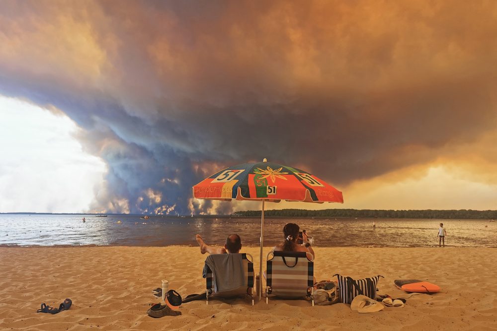 Strandgänger am Lacanau-See im Südwesten von Frankreich, sie sitzen unter einem Sonnenschirm und gegenüber von ihnen, am anderen Seeufer, steigt eine riesige Qualmwolke in die Luft