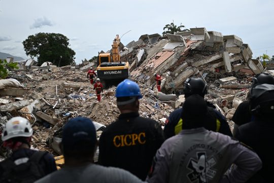 Freiwillige und Rettungssanitäter stehen still da, während Rettungskräfte Schalltests durchführen, um Überlebende aufzuspüren. Im Hintergrund sind Trümmer und ein Bagger zu sehen