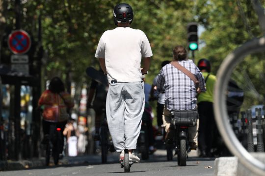 Die Straßenszene zeigt einen Scooterfahrer mit Helm, vor ih mein Fahrradfahrer ohne