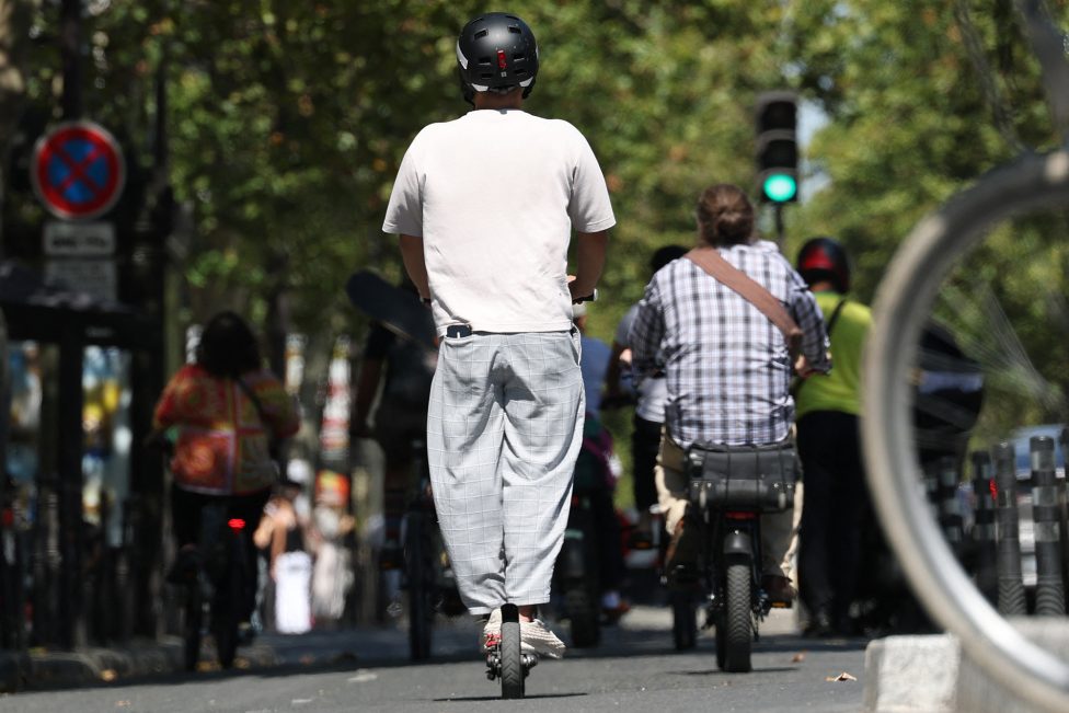 Die Straßenszene zeigt einen Scooterfahrer mit Helm, vor ih mein Fahrradfahrer ohne