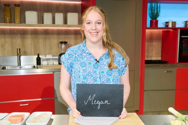 "Die Küchenschlacht": Kandidatin Megan Kohnen steht in der Küche der "Küchenschlacht" und hält ein Schild mit ihrem Namen in den Händen, dabei lächelt sie in die Kamera.