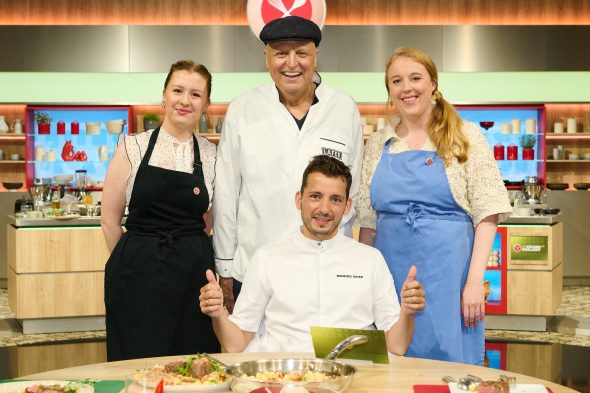 "Die Küchenschlacht", Sendung vom 28.08.2026: Maja Kunz, Johann Lafer, und Megan Kohnen stehen in der Studioküche vor dem Tisch, an dem Maurizio Oster mit nach oben gestreckten Daumen sitzt.