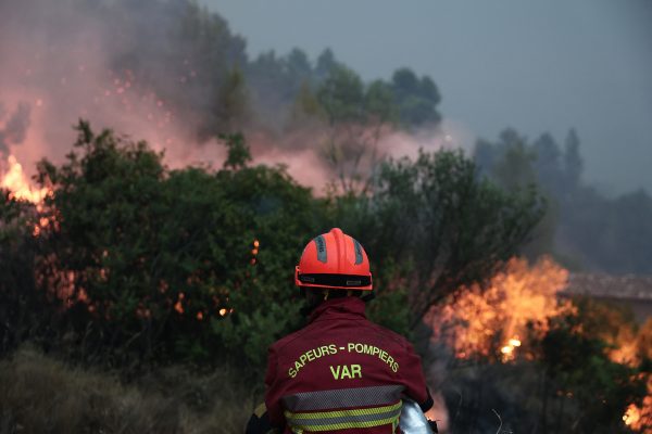 Feuerwehrmann mit dem Schriftzug "Var" auf dem Rücken, im Hintergrund sieht man den Waldbrand