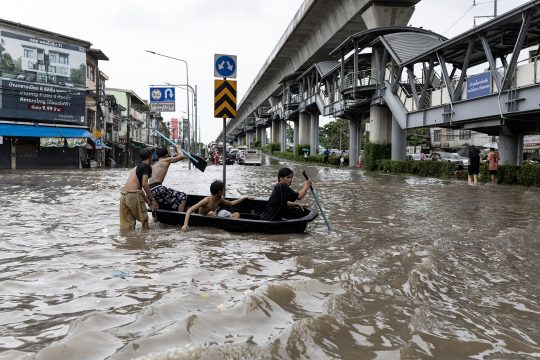 Überschwemmte Straße in Bangkok, junge Männer bewegen sich mithilfe eines Bootes fort
