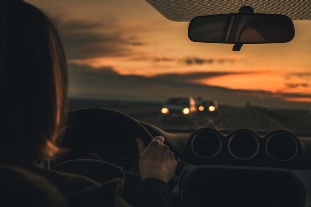 Eine Frau steuert eine Auto in der Dämmerung, ihr kommen zwei Autos mit eingeschalteten Scheinwerfern entgegen