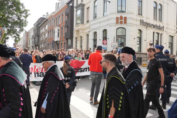 Im Vordergrund Angehörige des Lehrpersonals der Uni bei ihrem Umzug, im Hintergrund Menschen, die mit Banner in der Hand demonstrieren