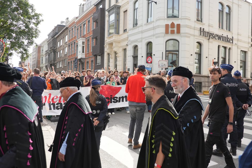 Im Vordergrund Angehörige des Lehrpersonals der Uni bei ihrem Umzug, im Hintergrund Menschen, die mit Banner in der Hand demonstrieren