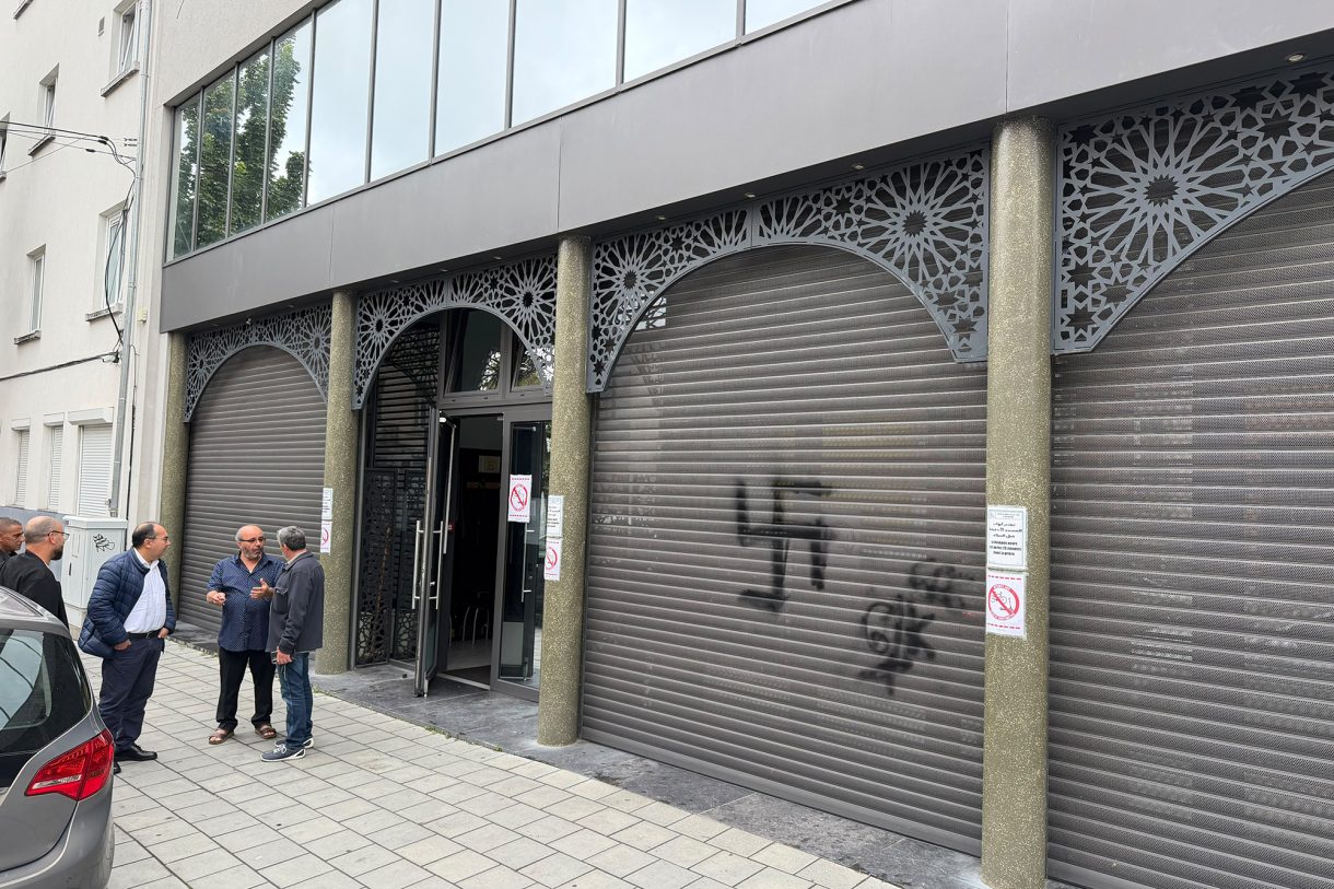 Das Bild zeigt die Front der Moschee in Laeken, auf einem der Rollläden ist ein augesprühtes chwarzes Hakenkreuz zu sehen. Einige Männer sehen vor dem Gebäude und reden miteinander