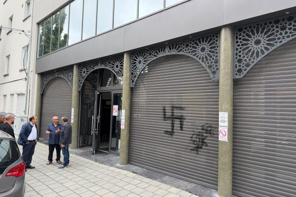 Das Bild zeigt die Front der Moschee in Laeken, auf einem der Rollläden ist ein augesprühtes chwarzes Hakenkreuz zu sehen. Einige Männer sehen vor dem Gebäude und reden miteinander