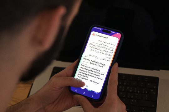 EinMann hält ein Smartphone in der hand, darauf ist eine Warnmeldung in arabischer und englische Sprache zu sehen