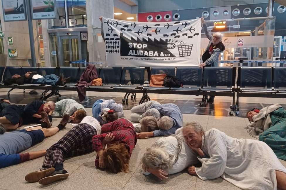 Menschen liegen im Flughafengebäude auf dem Boden und tun so, als würden sie schlafen - im Hintergrund wird ein Banner hochgehalten von der Protestgruppe "Stop Alibaba & Co"