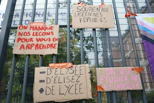 Pappschilder hängen an einem Schultor - auf ihnen steht unter anderem geschrieben: „Wir versäumen unseren Unterricht, um euch zu unterrichten“, „Weniger Élysée, weniger Gewählte, mehr Gymnasien“ und „Wir wollen lernen, statt Schulden anzuhäufen“