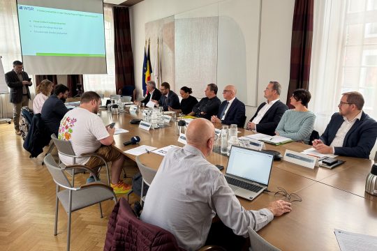 Vertreter von Regierung, WSR und Presse sitzen am Konferenztisch und blicken auf eine Leinwand