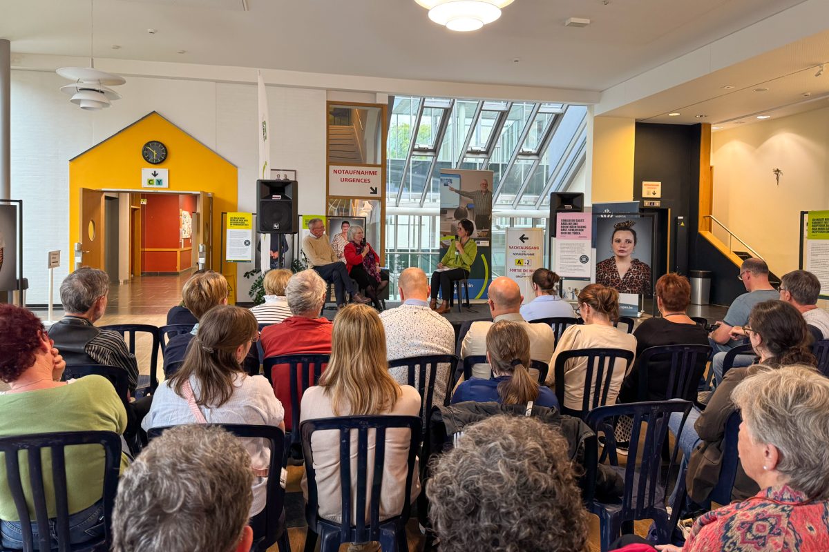 Ausstellungsbesucher sitzen auf Stühlen und hören bei einer Gesprächsrunde zu, die auf einem Podium zwischen drei Menschen stattfindet