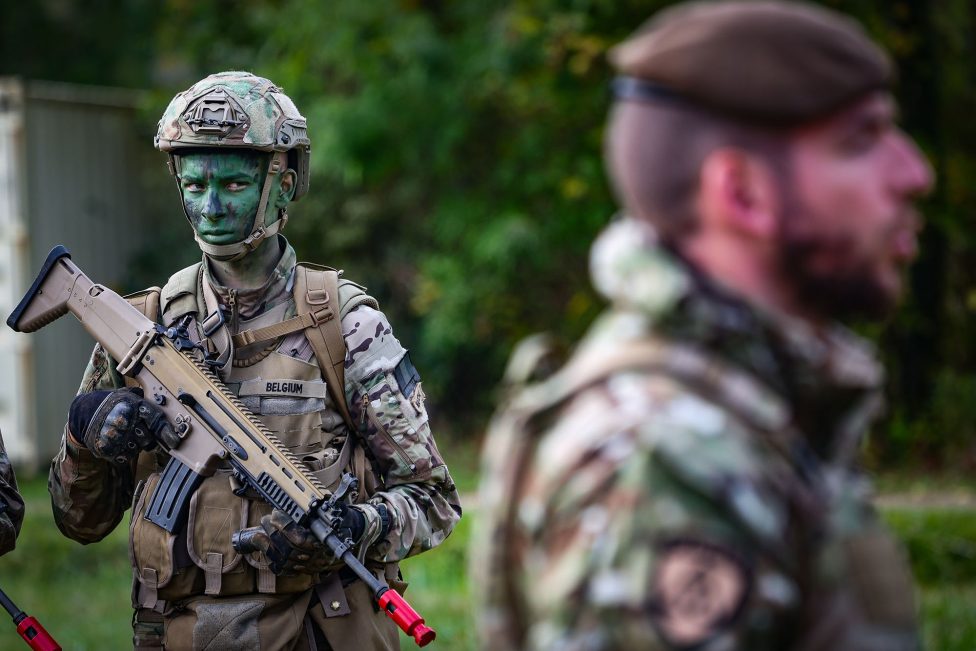 ein junger Mann in Tarnkleidung, mit Helm und Waffe, schaut auf einen Ausbilder