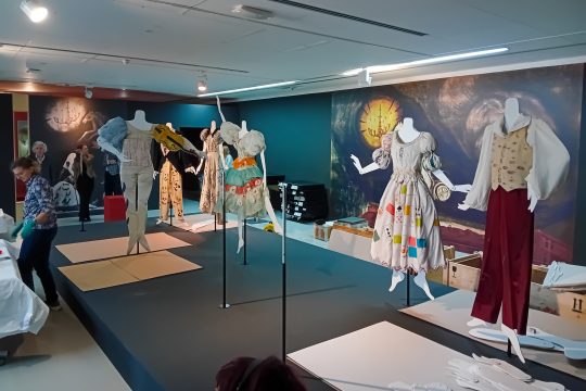 In einem Raum stehen Modepuppen, die unterschiedliche Kleidungsstücke tragen, die an Kunst von Marc Chagall erinnern - an den Wänden hängen große Abdrücke von gemalten Werken