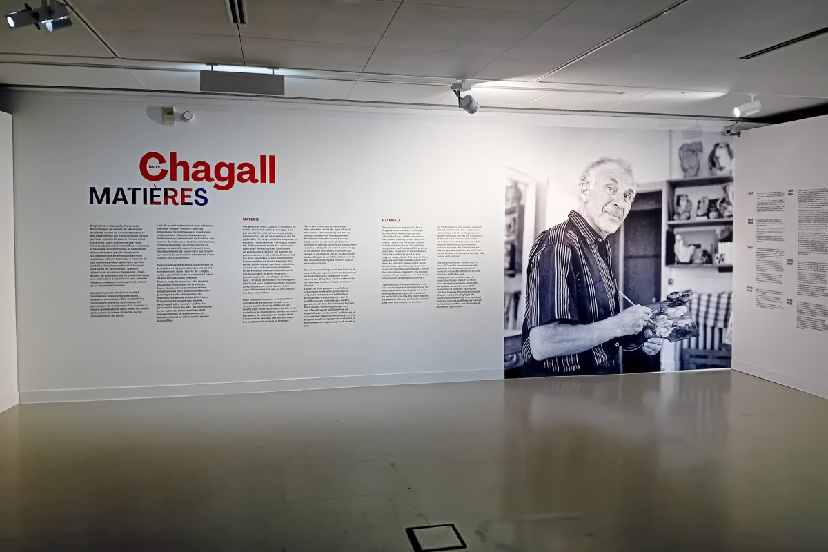 Auf einer Wand ist ein Schwarz-Weiß-Foto von Marc Chagall angebracht, das ihn beim Malen zeigt mit Pinsel und Palette in den Händen - daneben stehen Texte mit Informationen zur Ausstellung