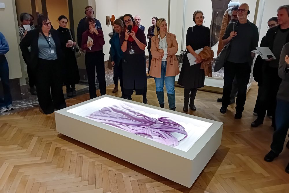 Besucher stehen um ein Exponat herum: ein in einer Vitrine liegendem lila Kleid