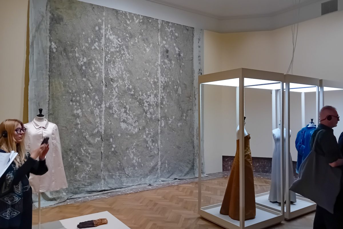 Kleider werden in Vitrinen gezeigt, an der Wand hängt ein großes Exponat aus grauem, beflecktem Stoff
