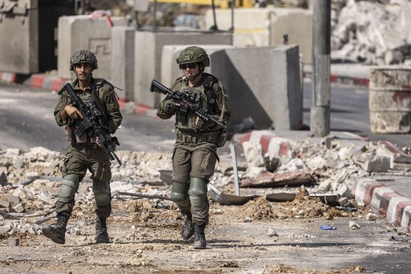 Zwei Israelische Soldaten in Tarn-Uniform patrouillieren auf der Straße, im Hintergrund Betonblocks einer Straßensperre, davor liegen Trümmer