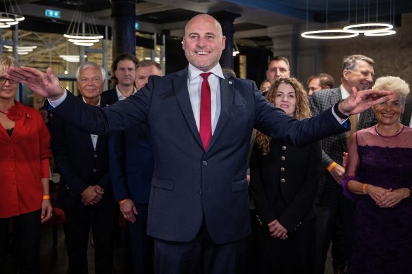 Der lettische Premierminister Andris Kulbergs breitet erfreut die Hände aus, hinter ihm stehen Mitglieder seiner Partei und blicken ebenfalls erfreut