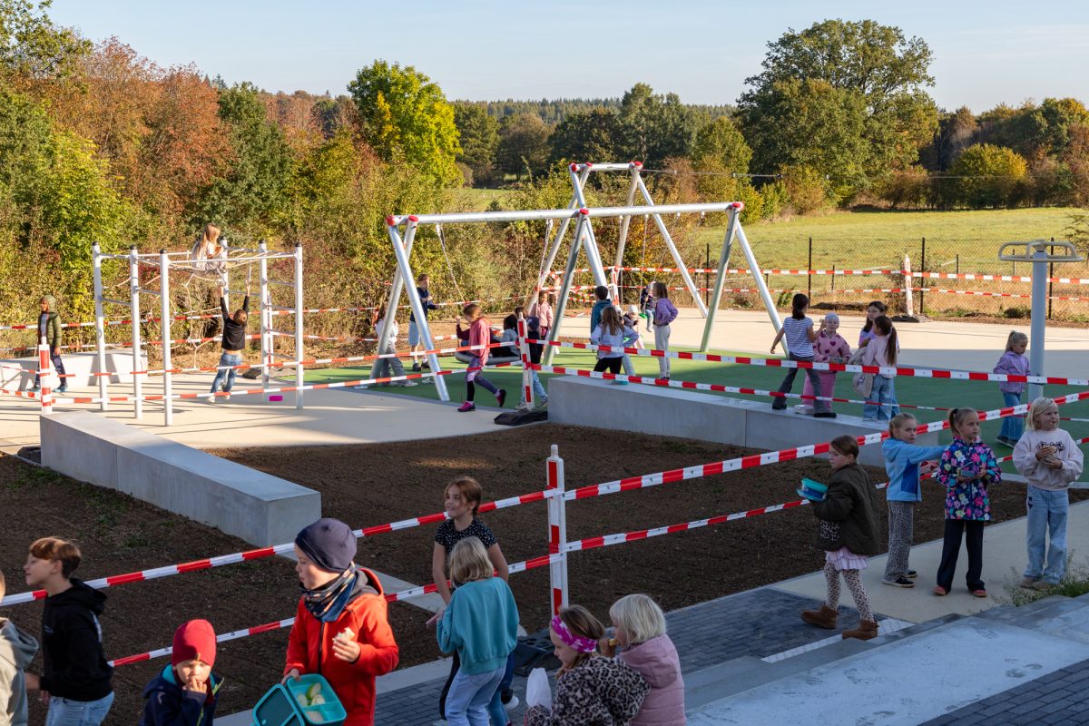 Kinder stehen auf dem Pausenhof, einige benutzen Spielgeräte, zum Beispiel eine Schaukel - in der Mitte ist ein Bereich mit Flatterband abgesperrt, der von Erde bedeckt ist