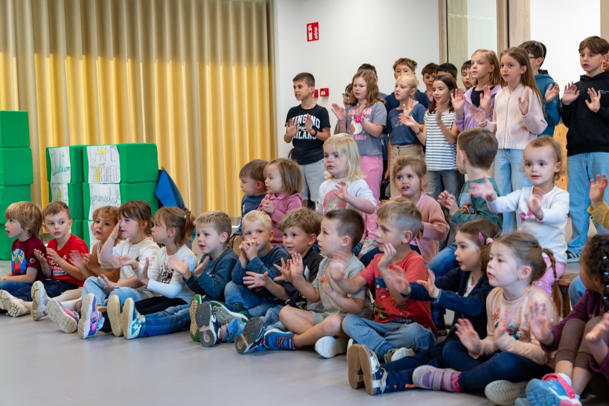 Kinder stehen zusammen, vor ihnen sitzen Kinder auf dem Boden - sie halten ihre Hände nach vorne - es scheint, als würden sie ein Lied singen