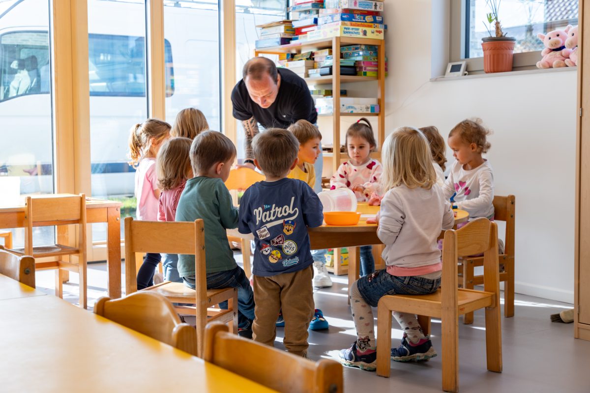 Kleine Kinder sitzen auf kleinen Stühlen an einem kleinen Tisch im Klassenraum, ein erwachsener Mann steht bei ihnen
