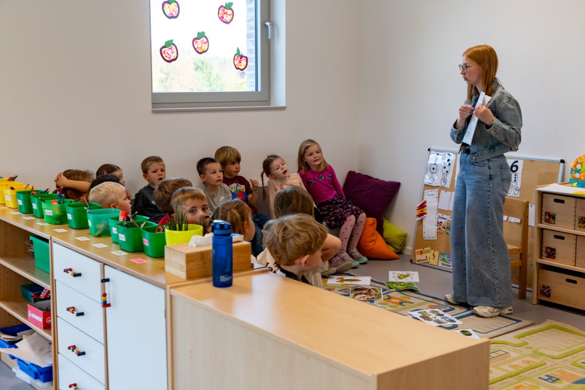 Lehrerin steht in einer Ecke des Raumes und hält einen Zettel in der Hand - die Kinder sitzen vor ihr auf dem Boden und interagieren mit ihr