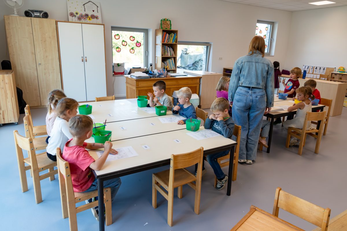 Kleine Kinder sitzen an kleinen Tischen im Klassenraum und malen, eine Lehrerin steht vor einem der Tische